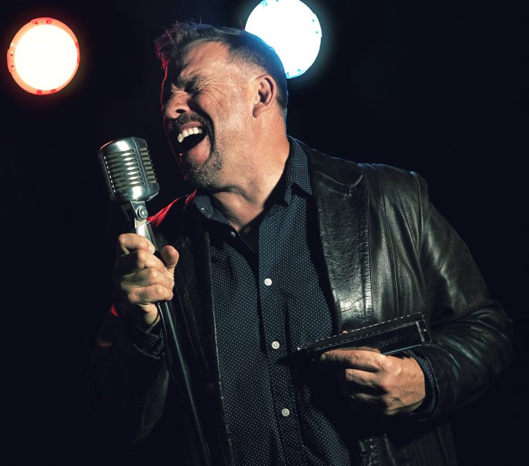 Chris O'Leary sings vocals live on stage while holding a harmonica.